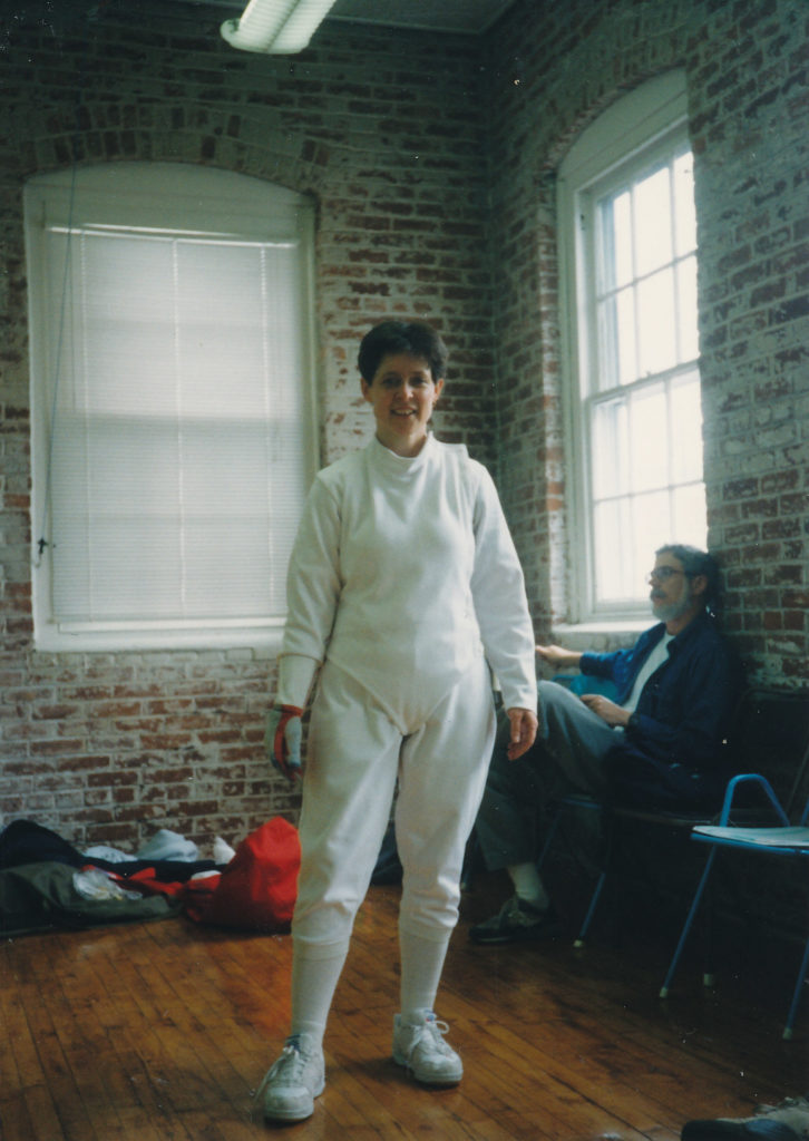 Delia standing inside a fencing club with brick interior walls and big windows. I am wearing full fencing whites. 