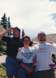Two men flank a woman on a hillside. The woman is wearing a US Olympic Training Center T-shirt and smiling, and the man on the right side has his arm around her.
