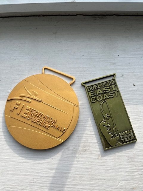 A big round gold medal reading "FIE: Federational Internationale d'Escrime" and a rectangular one reading "Dueal of the East Coast: Farrag Fencing Team."